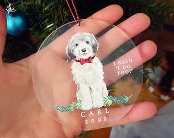 Könnte beinhalten: Ein runder, transparenter Acryl-Weihnachtsbaumschmuck mit einem weiß-grauen Hund, der eine rote Fliege trägt. Der Text lautet I SAID I DO, TOO! über dem Namen CARL und dem Jahr 2022. Grüne Tannenzweige sind am unteren Rand abgebildet.