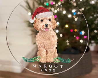 Può includere: Una decorazione natalizia rotonda in acrilico trasparente con un'illustrazione ad acquerello di un cane marrone chiaro dal pelo riccio che indossa un cappello da Babbo Natale rosso. Sotto il cane, rami di pino verdi e il testo bianco MARGOT 2022.