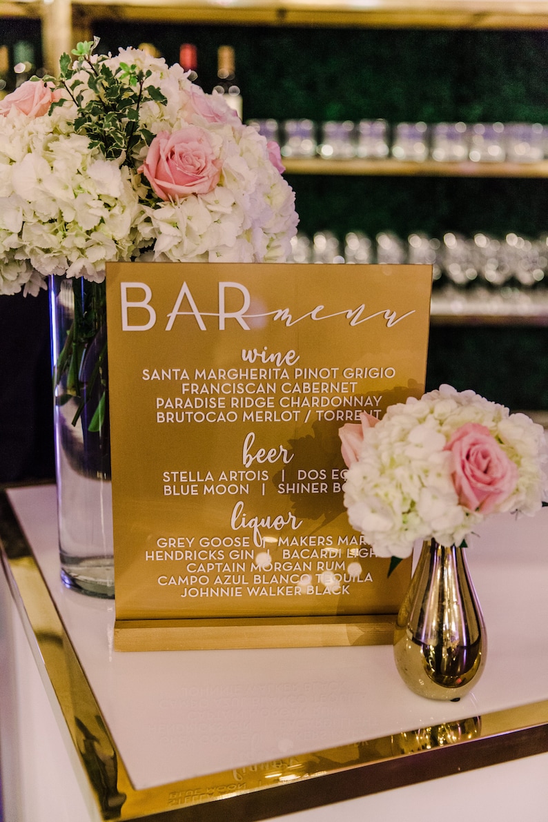 May include: A gold bar menu sign with white text listing wine, beer, and liquor options. The sign is on a white surface with a gold trim. There are white and pink flowers in vases on either side of the sign.