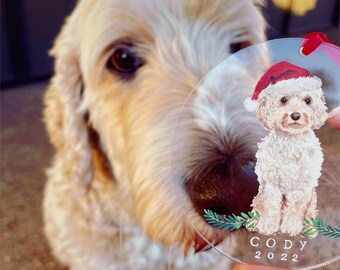 May include: A clear circular acrylic ornament featuring a watercolor illustration of a cream-colored dog wearing a red Santa hat. The dog sits above green pine sprigs. The text CODY and 2022 is printed in white at the bottom of the ornament.