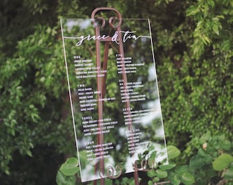 May include: A clear acrylic wedding seating chart with white calligraphy listing guest names and table numbers. The chart is on a rustic metal easel.