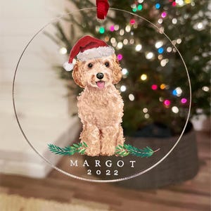 以下が含まれることがあります： サンタ帽をかぶったプードルのクリアな丸いクリスマスオーナメント。オーナメントには「MARGOT」という名前と「2022」の年がプリントされています。吊り下げ用の赤いリボンが付いています。背景にはクリスマスツリーがあります。