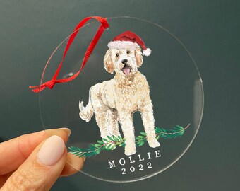 Może przedstawiać: Okrągła, przezroczysta ozdoba akrylowa z akwarelową ilustracją kremowego psa w czerwono-białej czapce Mikołaja. Pies stoi nad zielonymi gałązkami sosny. Poniżej nadrukowano napis MOLLIE 2022. Dołączona jest czerwona wstążka.