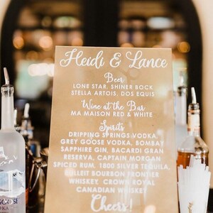 May include: A brown sign with white text listing the drinks available at a wedding reception for Heidi & Lance. The sign lists beer, wine, and spirits. The sign is on a gold stand.