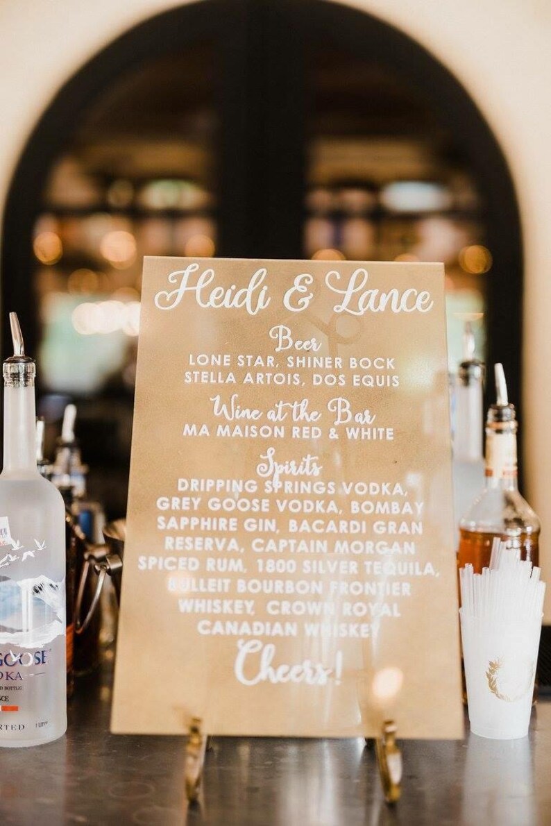 May include: A brown sign with white text listing the drinks available at a wedding reception for Heidi & Lance. The sign lists beer, wine, and spirits. The sign is on a gold stand.