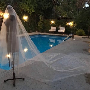 May include: A white tulle wedding veil displayed on a mannequin against a backdrop of a swimming pool and lush greenery.