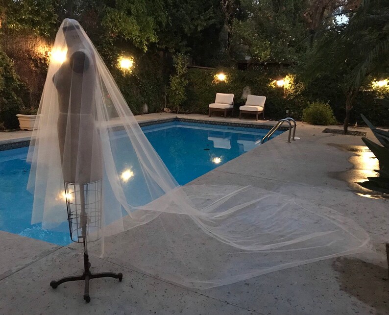 May include: A white tulle wedding veil displayed on a mannequin against a backdrop of a swimming pool and lush greenery.