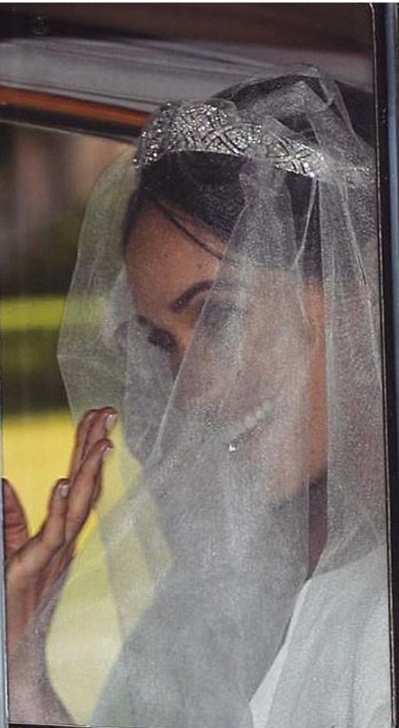 May include: A bride wearing a white veil and a tiara with a diamond design. The veil is sheer and covers her face. She is wearing a white dress and has her hand raised.