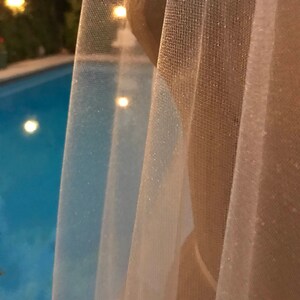 May include: A sheer white curtain with a subtle shimmer, partially obscuring a view of a swimming pool with blue water and lights.