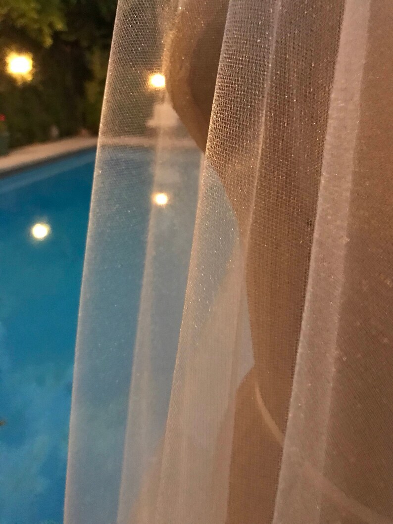 May include: A sheer white curtain with a subtle shimmer, partially obscuring a view of a swimming pool with blue water and lights.
