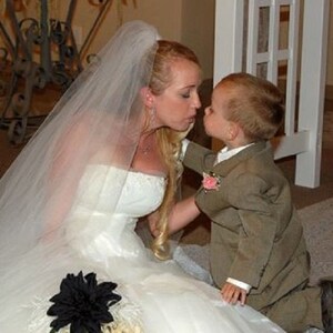May include: A bride in a white wedding dress with a veil kisses a young boy in a brown suit. The boy is wearing a pink flower on his lapel.