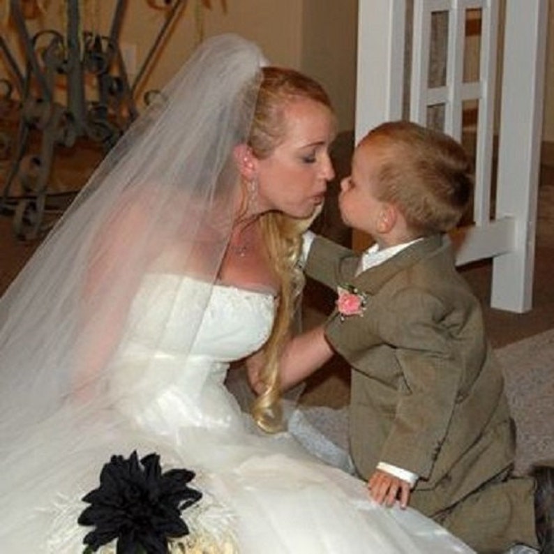 May include: A bride in a white wedding dress with a veil kisses a young boy in a brown suit. The boy is wearing a pink flower on his lapel.