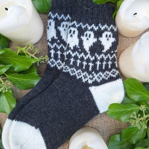 May include: A pair of gray and white knit socks with a ghost pattern. The socks are folded and displayed on a burlap surface with white candles and green leaves.