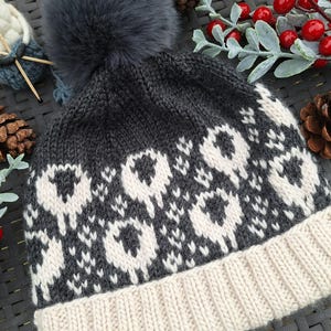 May include: A grey and white knitted hat with a black and white sheep pattern. The hat has a fluffy grey pom pom on top and a white brim.