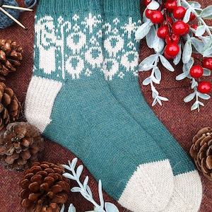 May include: A pair of teal knitted socks with a white pattern of stylized figures and floral designs. The heel and toe are light beige. The socks are surrounded by pine cones and red berries.