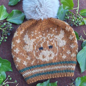 Puede incluir: Un gorro de punto marrón con un diseño de reno de color beige claro y un pompón gris claro y esponjoso. El gorro presenta una banda a rayas verdes y marrones en la base. Un accesorio de invierno.
