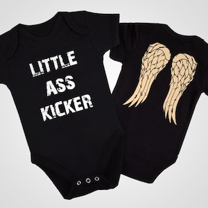 Peut inclure: Deux bodies bébé noirs. Un body a le texte "LITTLE ASS KICKER" imprimé en blanc. L'autre body a une paire d'ailes d'ange imprimées au dos en beige.
