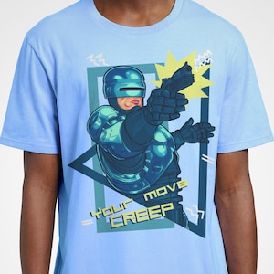 May include: Pale blue t-shirt with a graphic of a futuristic police officer aiming a gun. The design includes the text "YOUR MOVE CREEP" in a geometric frame. The shirt is a short-sleeved crew neck.