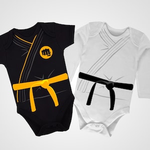 Puede incluir: Dos bodies para bebé, uno negro con un cinturón amarillo y un símbolo de puño, y otro blanco con un cinturón negro. Ambos bodies están diseñados para parecer uniformes de karate.