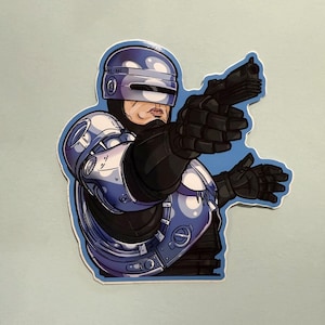 May include: A sticker featuring a stylized illustration of Robocop, a fictional cyborg police officer. The artwork depicts Robocop in his signature blue and silver armour, holding a black handgun and pointing it forward. The sticker has a blue outline.