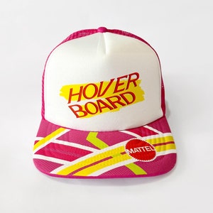 Pode incluir: Um chapéu de caminhoneiro branco e rosa com um logotipo "Hover Board" amarelo e vermelho e um logotipo Mattel vermelho.