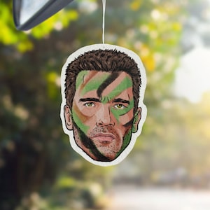 May include: A white car air freshener featuring a cartoon image of a man's face with green and brown camouflage paint.