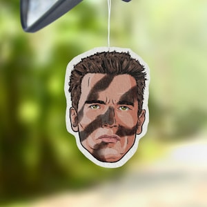 May include: A white car air freshener featuring a cartoon illustration of a man's face with black and brown camouflage stripes across the face.