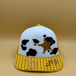 Puede incluir: Una gorra tipo trucker blanca con estampado de manchas de vaca negras, una insignia de sheriff dorada en forma de estrella y una visera amarilla con cuadrícula roja. La palabra ANDY está escrita en negro en la visera, con un pequeño lazo blanco.