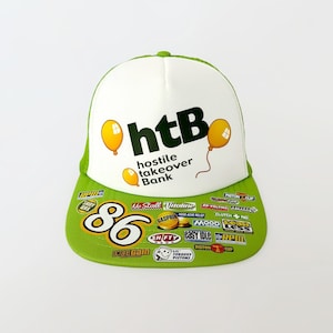 May include: A green and white trucker hat with the text "htB hostile takeover Bank" printed on the front. The hat has a variety of colourful patches and stickers on the brim, including "No Stall", "Vitutine", "Casprin", "Mood", "Clutch", "Easy Idle", "RPM", "Torquey Pistons", and "Piston".