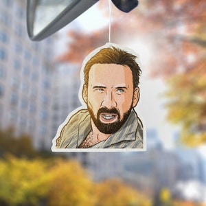 May include: A car air freshener featuring a cartoon illustration of a man with a beard and a serious expression. The man is wearing a light brown shirt with a striped pattern.