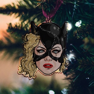 May include: A black and gold Christmas bauble depicting a woman wearing a black cat mask with a blonde wig. The bauble features a detailed illustration of the woman's face with a fierce expression.