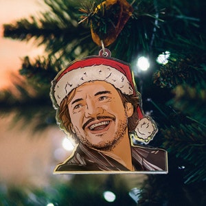May include: A Christmas ornament featuring a cartoon illustration of a man wearing a Santa hat and a scarf. The ornament is made of metal and has a gold finish.