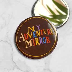 Può includere: Uno specchio compatto marrone con un design colorato che dice "MY ADVENTURE MIRROR".
