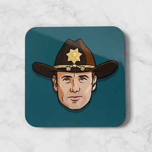 May include: Square coaster with a cartoon illustration of a man in a brown cowboy hat with a gold sheriff's badge. The man has blue eyes and a neutral expression, on a teal background.