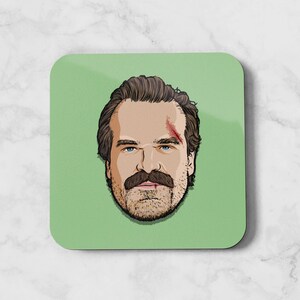 Puede incluir: Un posavasos cuadrado verde con una ilustración de dibujos animados de un hombre con bigote y un corte en la frente.