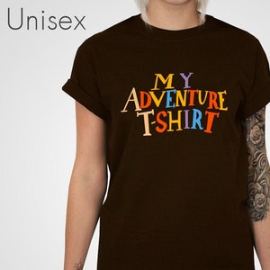Può includere: Una maglietta marrone con il testo "MY ADVENTURE T-SHIRT" in lettere colorate.