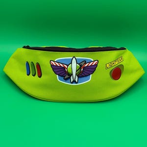 May include: A lime green fanny pack featuring a black zipper closure. The front displays three vertical oval buttons in blue, green, and red, a central space ranger emblem with purple wings, and a yellow rectangular patch with the text LIGHTYEAR.