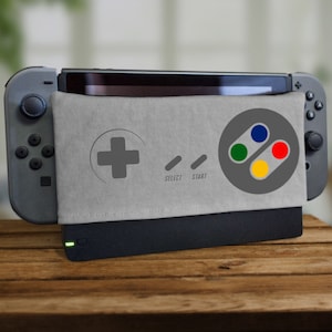 Puede incluir: Una consola Nintendo Switch gris con un diseño de mando Super Nintendo gris y blanco en la carcasa. La carcasa tiene las palabras "Select" y "Start" impresas en ella. La consola está sentada sobre una superficie de madera.
