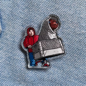 Puede incluir: Un pin de esmalte rojo y blanco con un niño montando una bicicleta con un extraterrestre en una cesta. El extraterrestre lleva una manta blanca y tiene ojos grandes.