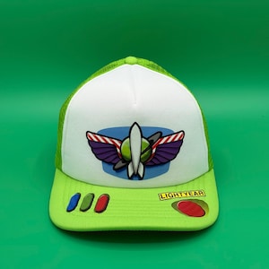 Puede incluir: Una gorra tipo trucker blanca y verde lima con un emblema de Space Ranger en el panel frontal. La visera es verde lima con tres pequeños botones ovalados en azul y rojo, y un parche rectangular amarillo con el texto LIGHTYEAR en negro.