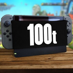 Puede incluir: Una consola Nintendo Switch negra con una funda negra que dice "100t" en letras blancas. La consola está sentada sobre una superficie de madera.