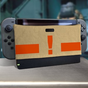 Puede incluir: Una funda beige para consola Nintendo Switch con rayas naranjas y un signo de exclamación. La funda está diseñada para parecerse a una señal de advertencia.