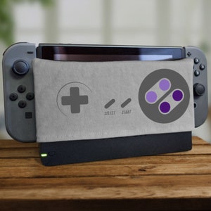Puede incluir: Una consola Nintendo Switch gris con un diseño de mando Super Nintendo gris y morado en la carcasa. La carcasa tiene las palabras "Select" y "Start" impresas.