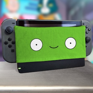 Könnte beinhalten: Eine grüne Nintendo Switch Konsolenhaut mit einem Comic-Gesicht. Das Gesicht hat zwei weiße Augen mit schwarzen Pupillen und ein schwarzes Lächeln.