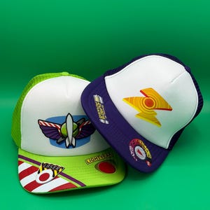 Puede incluir: Dos gorras tipo trucker con diseños de Buzz Lightyear y Zurg. La gorra izquierda es blanca y verde lima con un emblema de guardián espacial y texto láser. La derecha es blanca y morada con un emblema de rayo amarillo y un gráfico de dial circular.