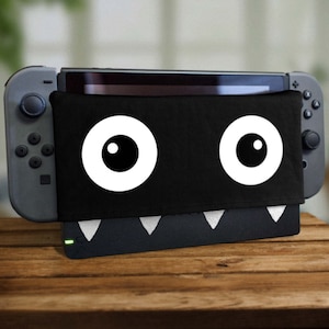 Peut inclure: Console de jeu Nintendo Switch noire avec un motif de visage de monstre noir et blanc sur le devant. Le monstre a deux grands yeux blancs avec des pupilles noires.