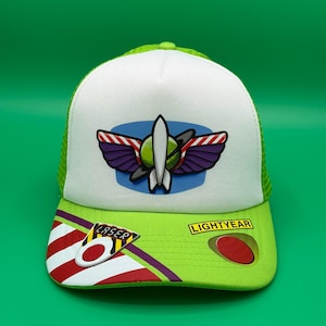 May include: A white and lime green trucker cap featuring a Space Ranger emblem on the front panel. The brim displays red and white diagonal stripes, a yellow rectangular patch with the text LIGHTYEAR, and a circular red accent with the word LASER.