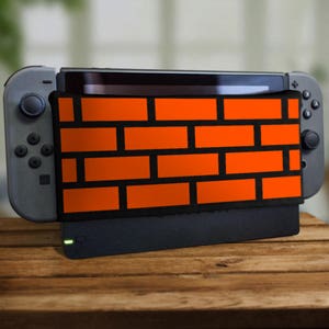 Peut inclure: Une console Nintendo Switch noire avec un motif de briques orange personnalisé sur l'écran. La console est dans un support et est éteinte.