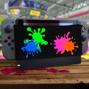 Könnte beinhalten: Eine schwarze Nintendo Switch Dock-Abdeckung mit bunten Farbspritzern in Grün, Blau, Pink und Orange. Die Abdeckung befindet sich auf einer grauen Nintendo Switch Dockingstation, mit angeschlossener Konsole. Der Hintergrund ist unscharf.