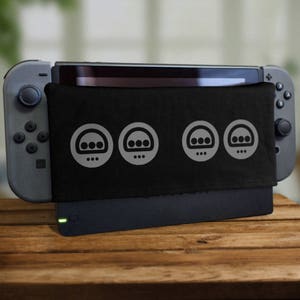 Puede incluir: Una consola Nintendo Switch negra con una cubierta de tela negra que presenta tres iconos grises. Los iconos son estilizados y se asemejan a un mando de videojuego.